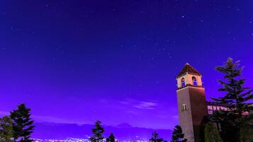Vista nocturna de una torre con un reloj, rodeada de árboles y un paisaje urbano al fondo.
