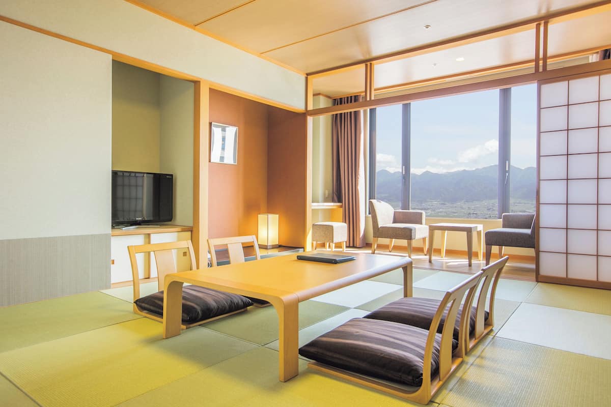 Ein modernes Hotelzimmer mit Tatami-Matten, einem Holzespeisebereich, Stühlen, einem Fernseher und einem großen Fenster mit Blick auf die Berge.