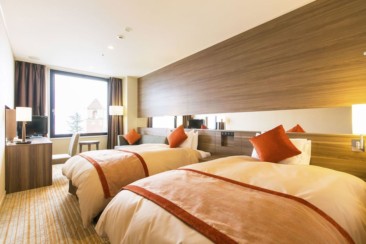 Ein Hotelzimmer mit zwei Betten, einem Schreibtisch mit Lampe, einem Fernseher und einem großen Fenster mit Vorhängen.