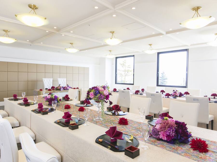 Un salón de conferencias con una larga mesa dispuesta para una reunión, sillas blancas y ventanas grandes.