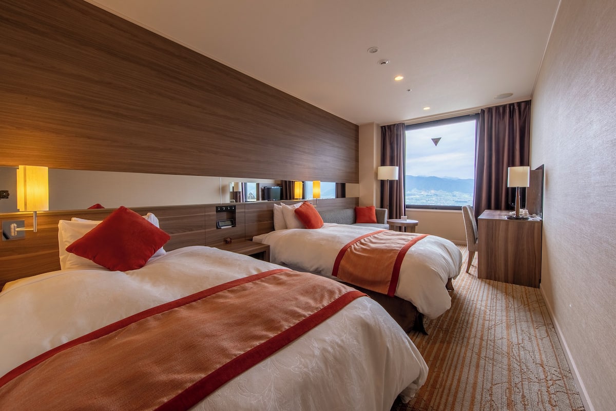 Ein Hotelzimmer mit zwei Betten, einem hölzernen Kopfteil, einem großen Fenster mit Aussicht, einem Schreibtisch mit Lampe und einem Fernseher.