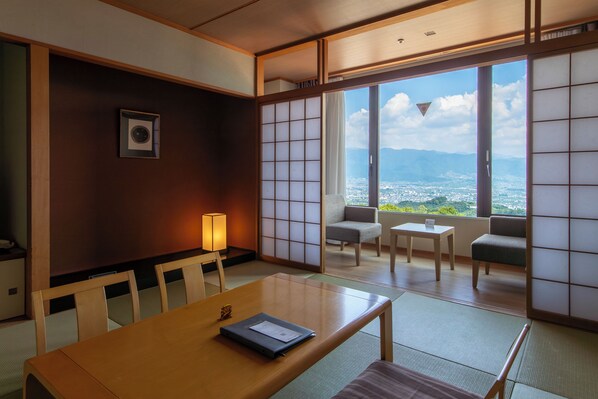 A traditional Japanese room with tatami flooring, a low table with a laptop, and a view of a cityscape through large sliding doors.