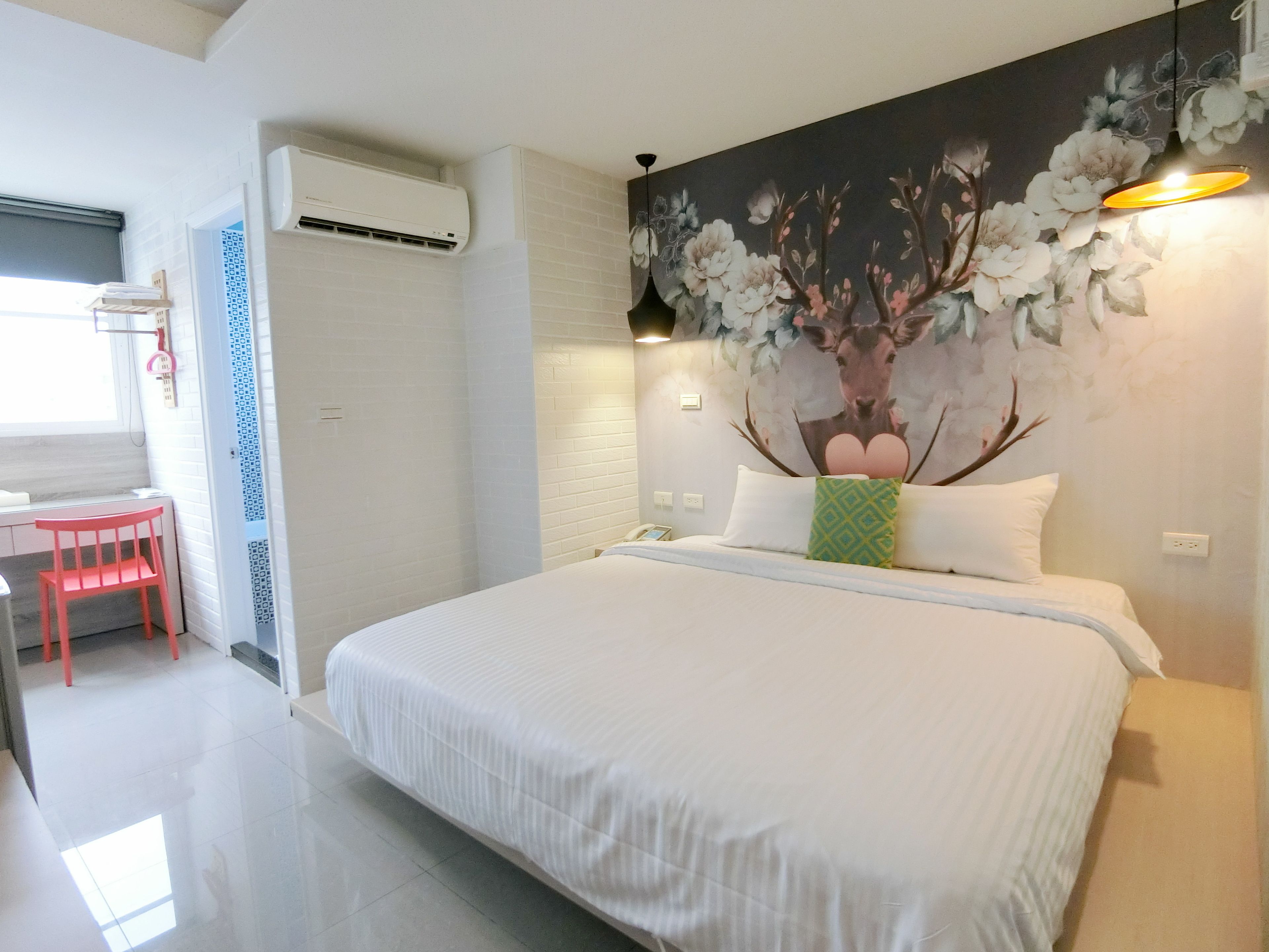 A bedroom with a large bed, a desk, and a chair. There is a wall-mounted air conditioner and a painting of flowers and a deer.