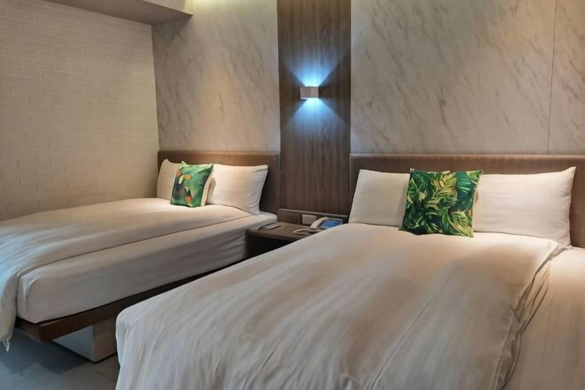 Habitación de hotel con dos camas, una mesita de noche, una lámpara fijada a la pared y un panel de madera.