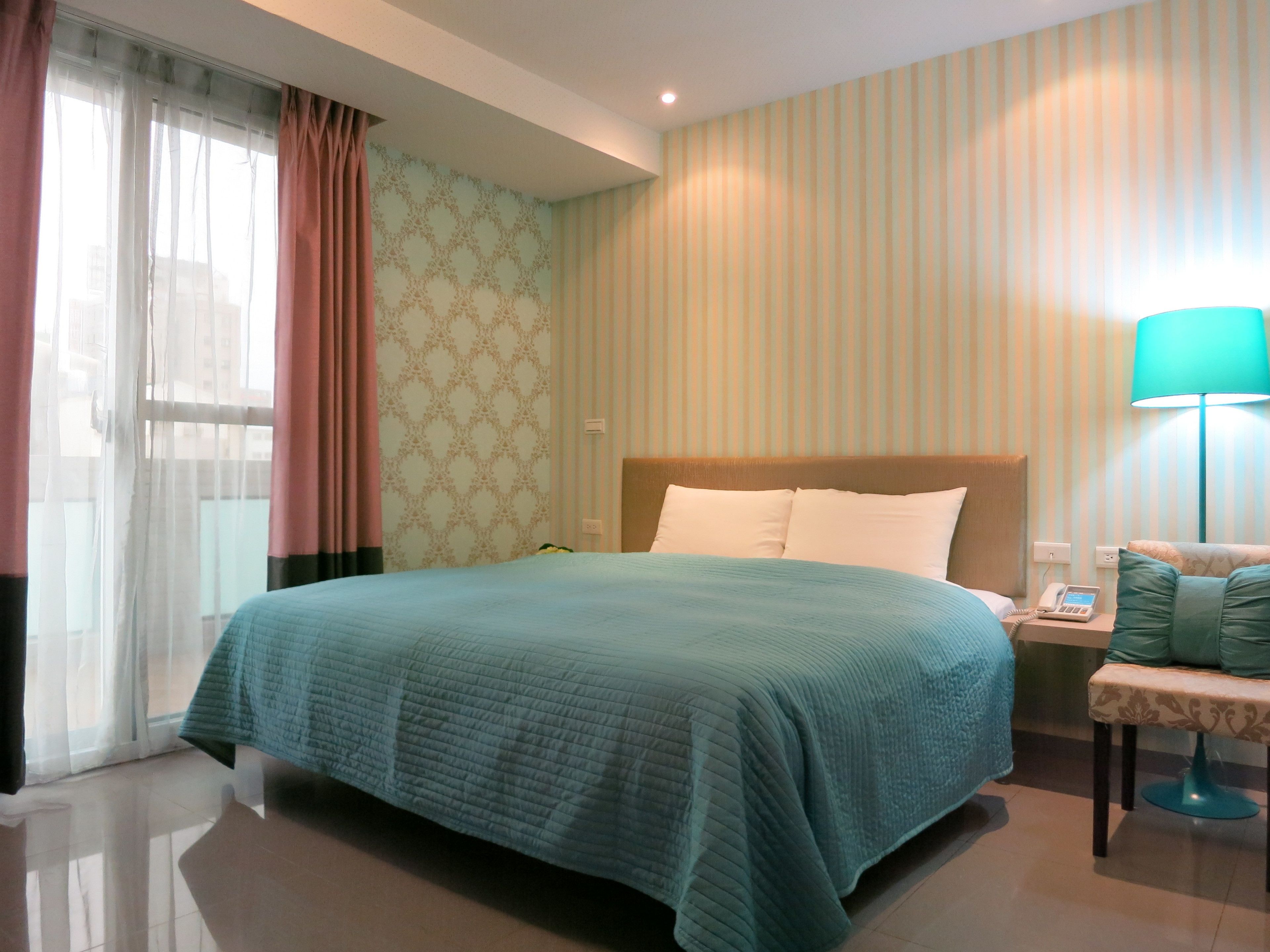 Una habitación de hotel con una cama grande, una mesita de noche con lámpara y una ventana con cortinas.