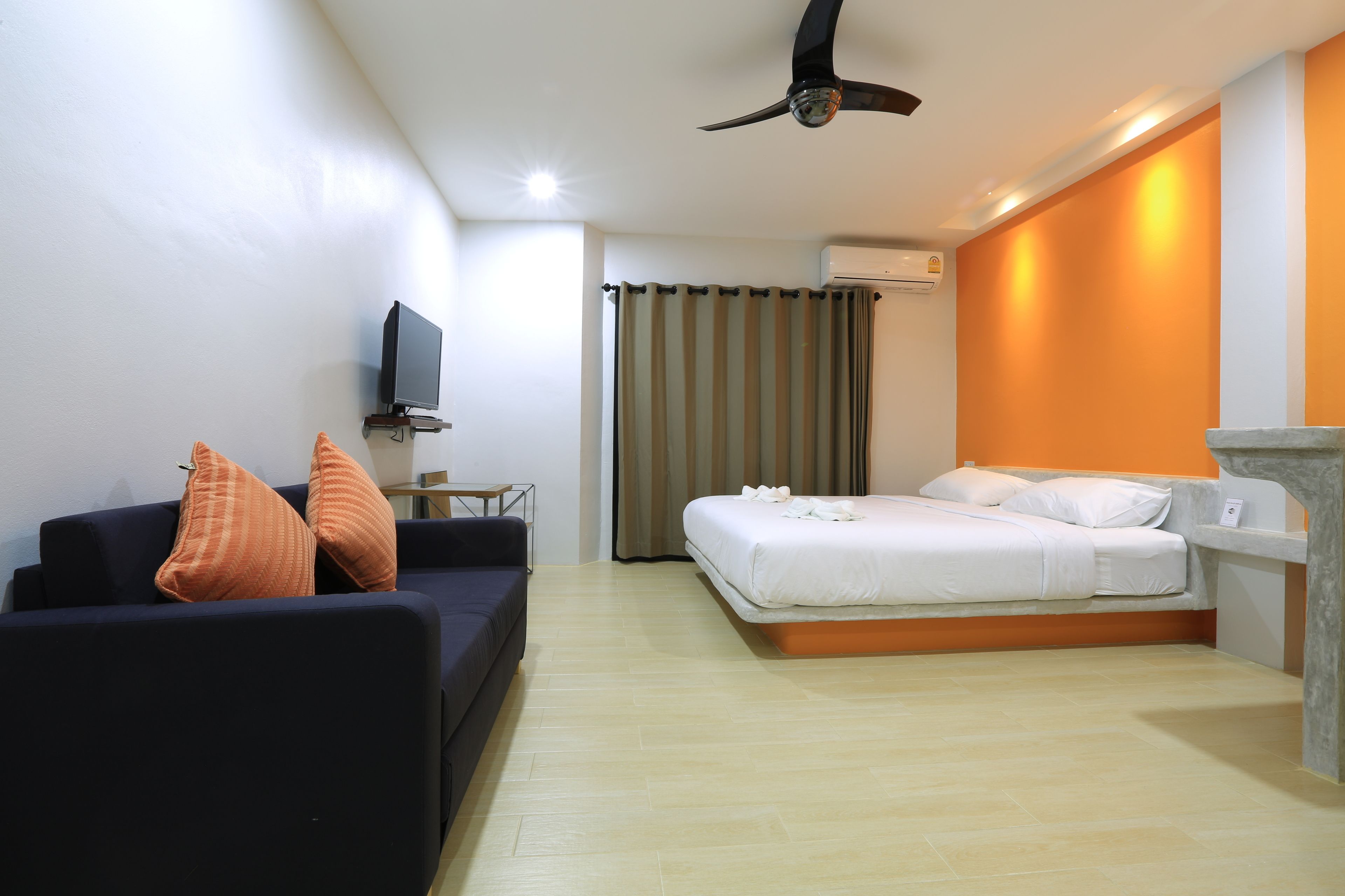 Una habitación de hotel con una cama, un sofá con almohadas naranjas, un ventilador de techo, un equipo de aire acondicionado y un televisor sobre un soporte.