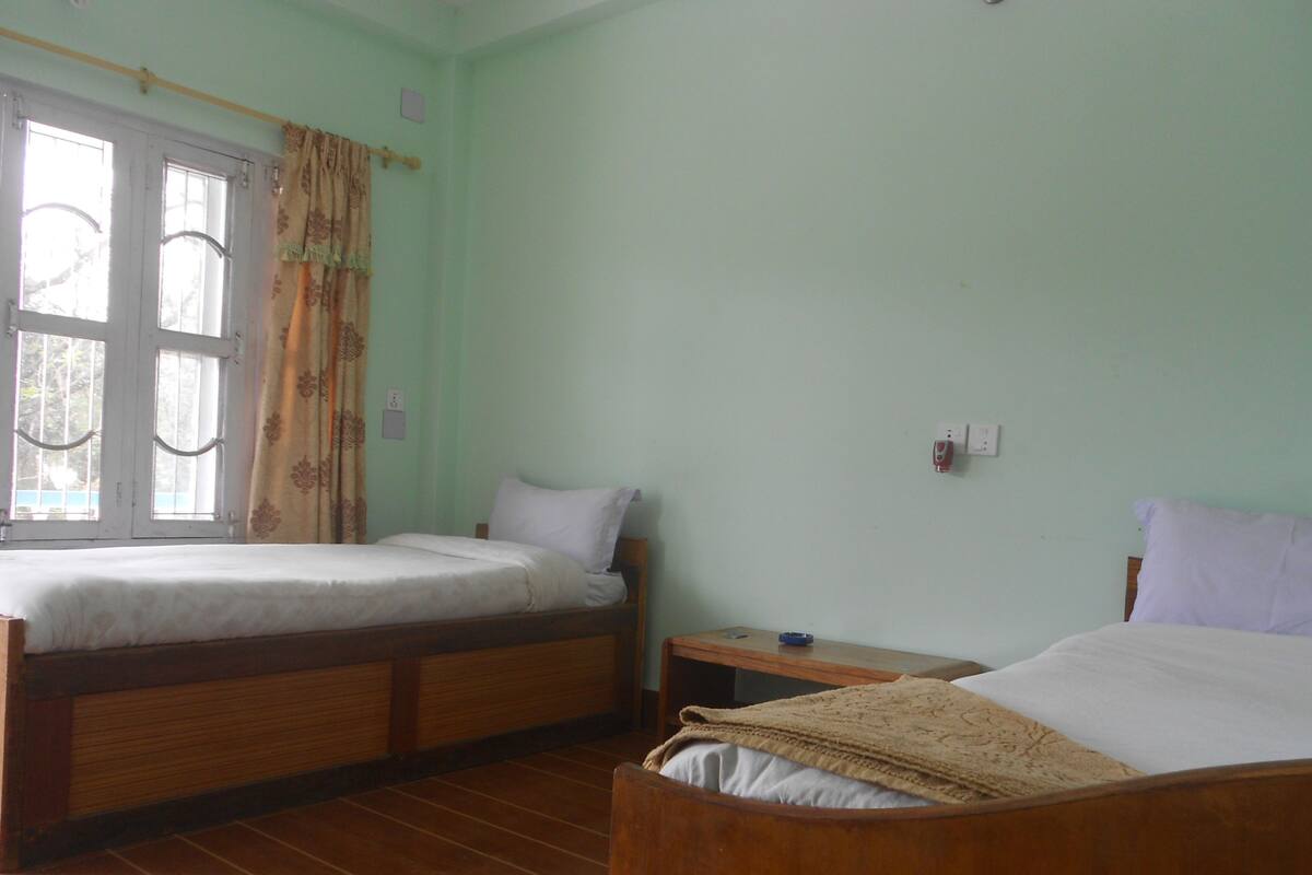 A room with two wooden beds, a window with curtains, a small wooden nightstand, and a wall socket.