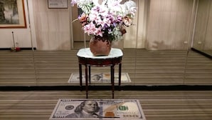 Une pièce avec un plancher en bois, une table blanche ornée d’un vase de fleurs et un billet d’un dollar sur le sol.