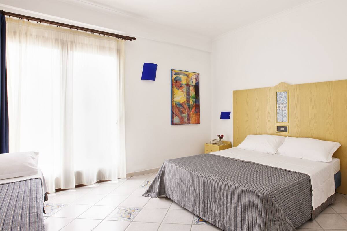 Une chambre d’hôtel avec un lit, une table de chevet, un tableau et une fenêtre avec des rideaux.