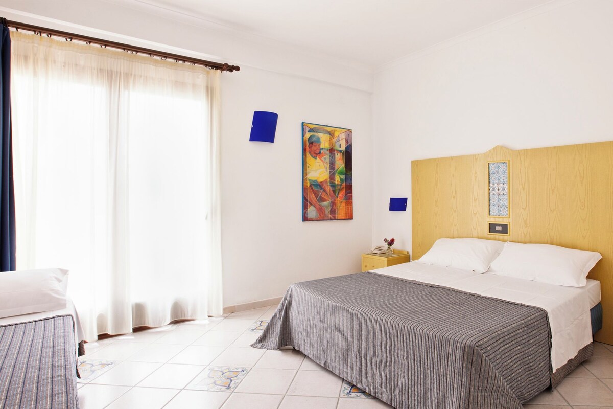 A hotel room with a bed, a nightstand, a painting, and a window with curtains.
