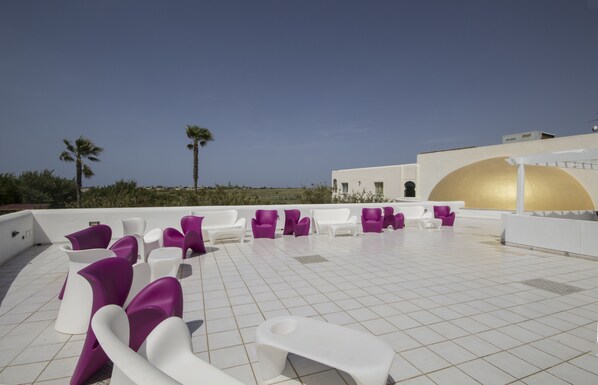 A rooftop terrace with white and purple seating, palm trees, and a white building with a golden arch.