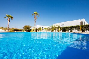 Une piscine d’un bleu éclatant, bordée de chaises longues blanches et entourée de palmiers, le tout adossé à un bâtiment blanc aux fenêtres en arc de cercle.