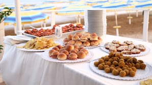 Une table de buffet garnie de divers plats et surmontée d’une pile d’assiettes, le tout installé sur une plage, avec des parasols en arrière-plan.