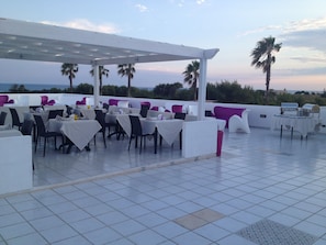 Un área de comedor al aire libre con sillas y mesas blancas y púrpuras, y una pérgola.