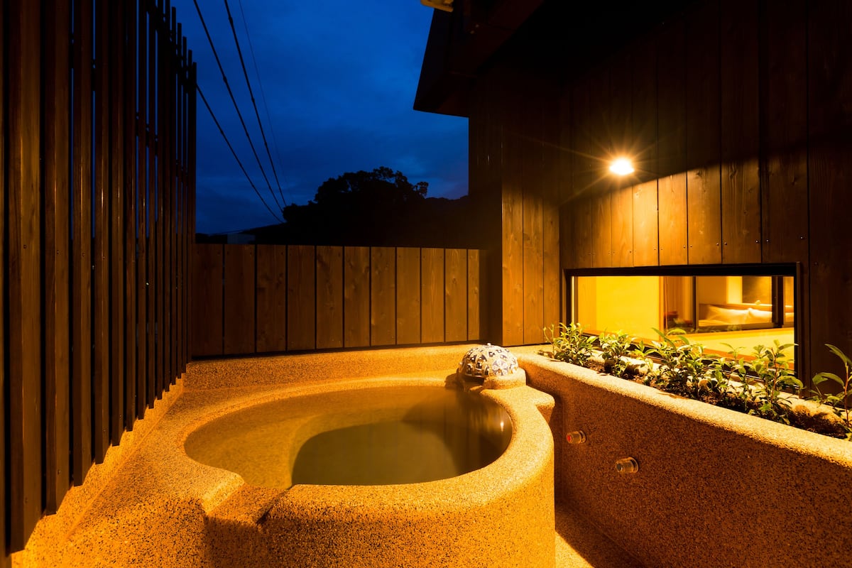 A hot tub in a wooden outdoor setting with a wooden fence and a window.