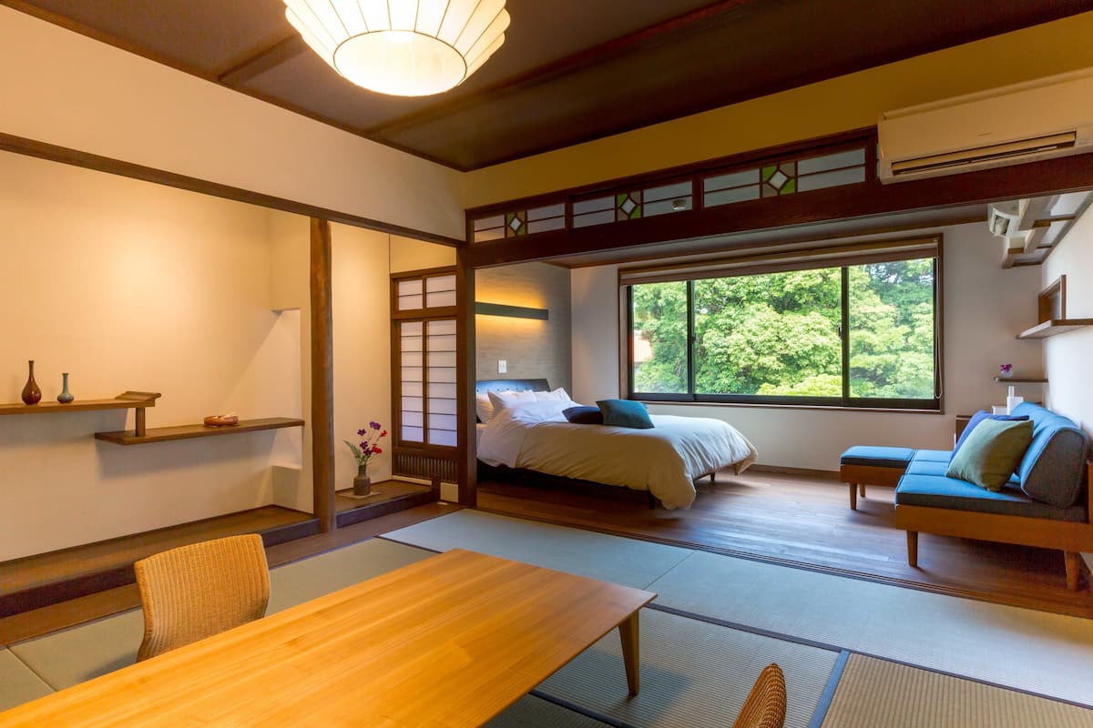A traditional Japanese-style room with tatami flooring, a low wooden table, a sofa, and a bed with a view of greenery outside.