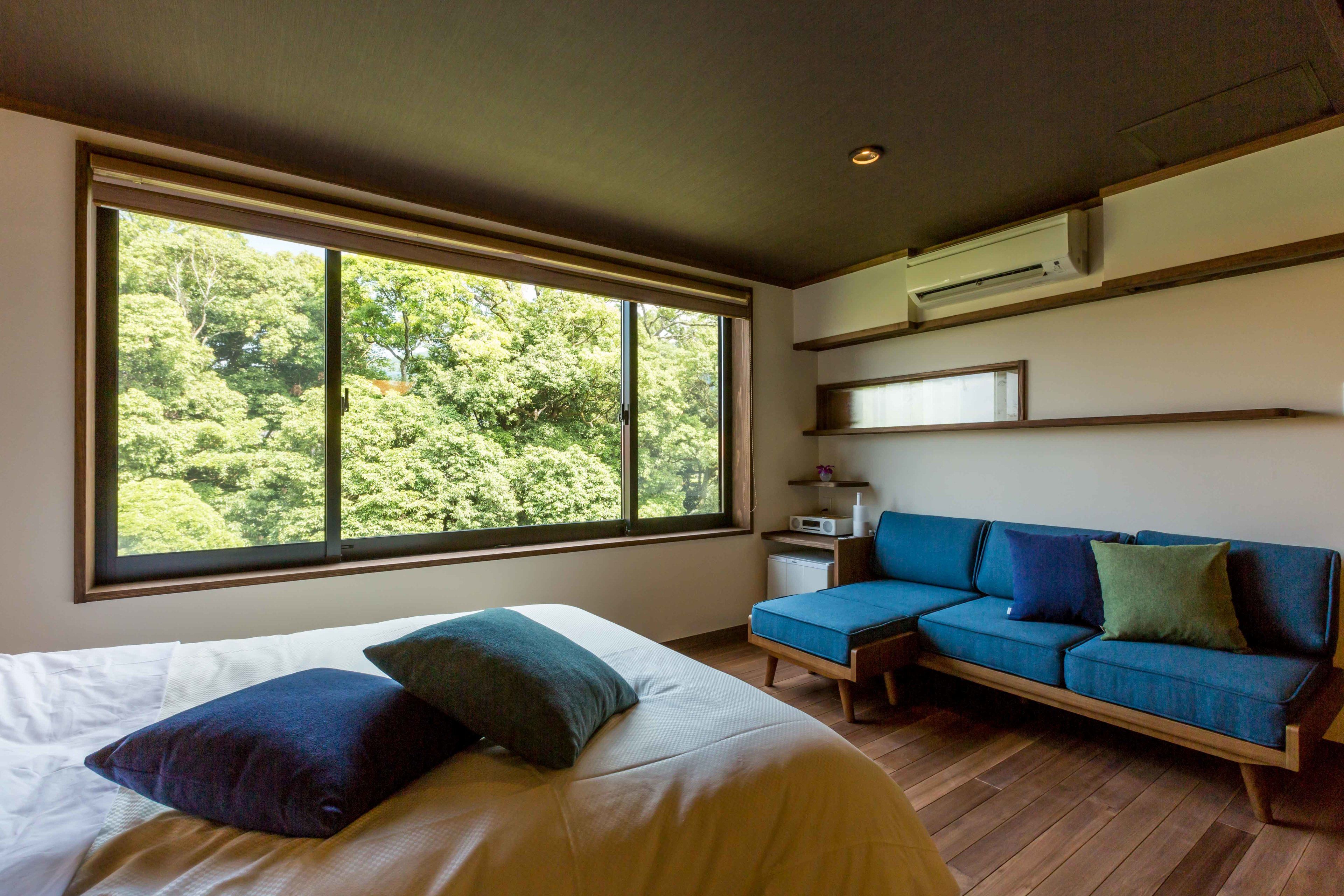 A bedroom with a large window, a bed, a blue sofa, and a small bedside table.