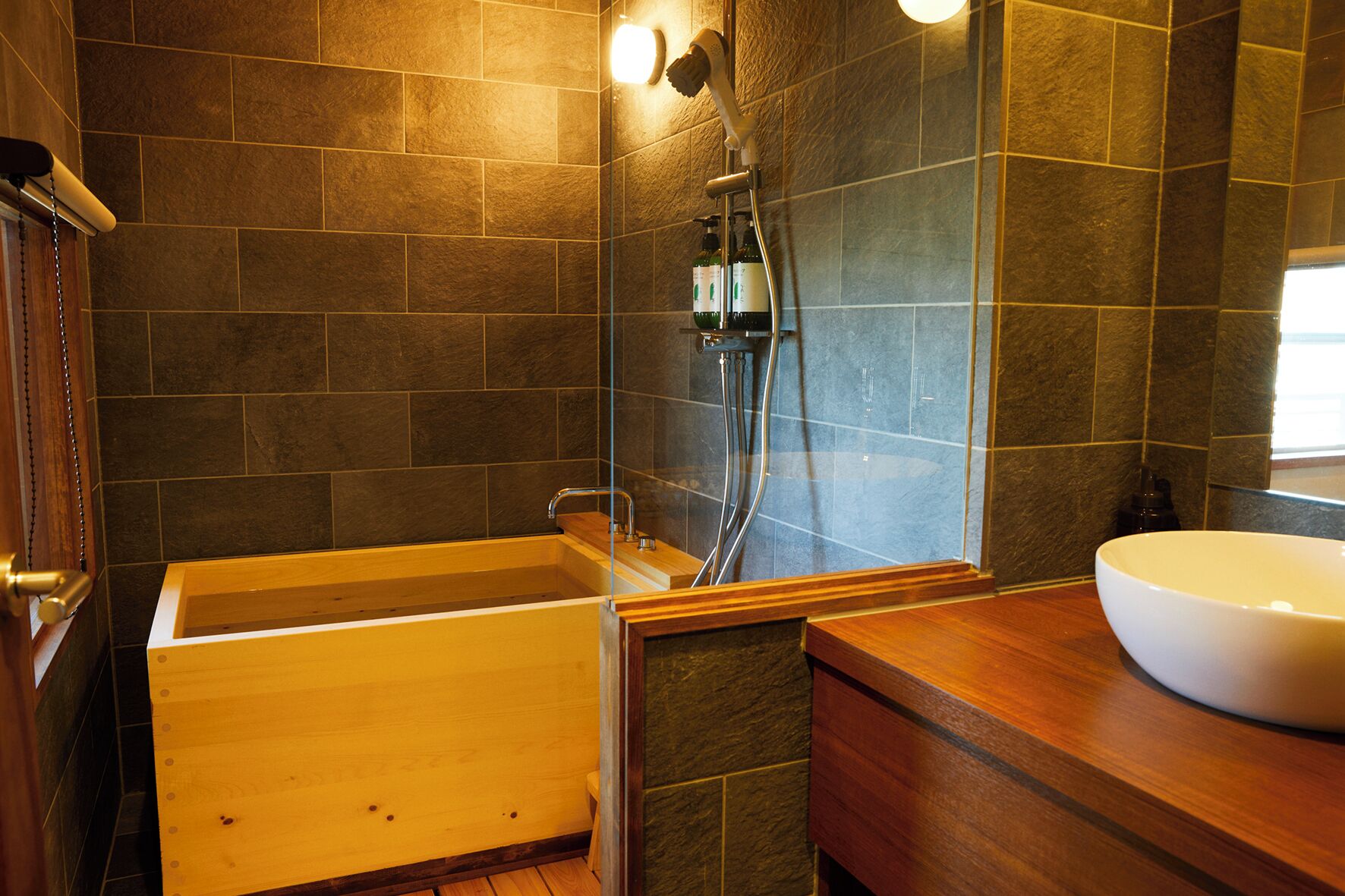 Un baño con bañera de madera, ducha con mampara de vidrio y lavamanos blanco.