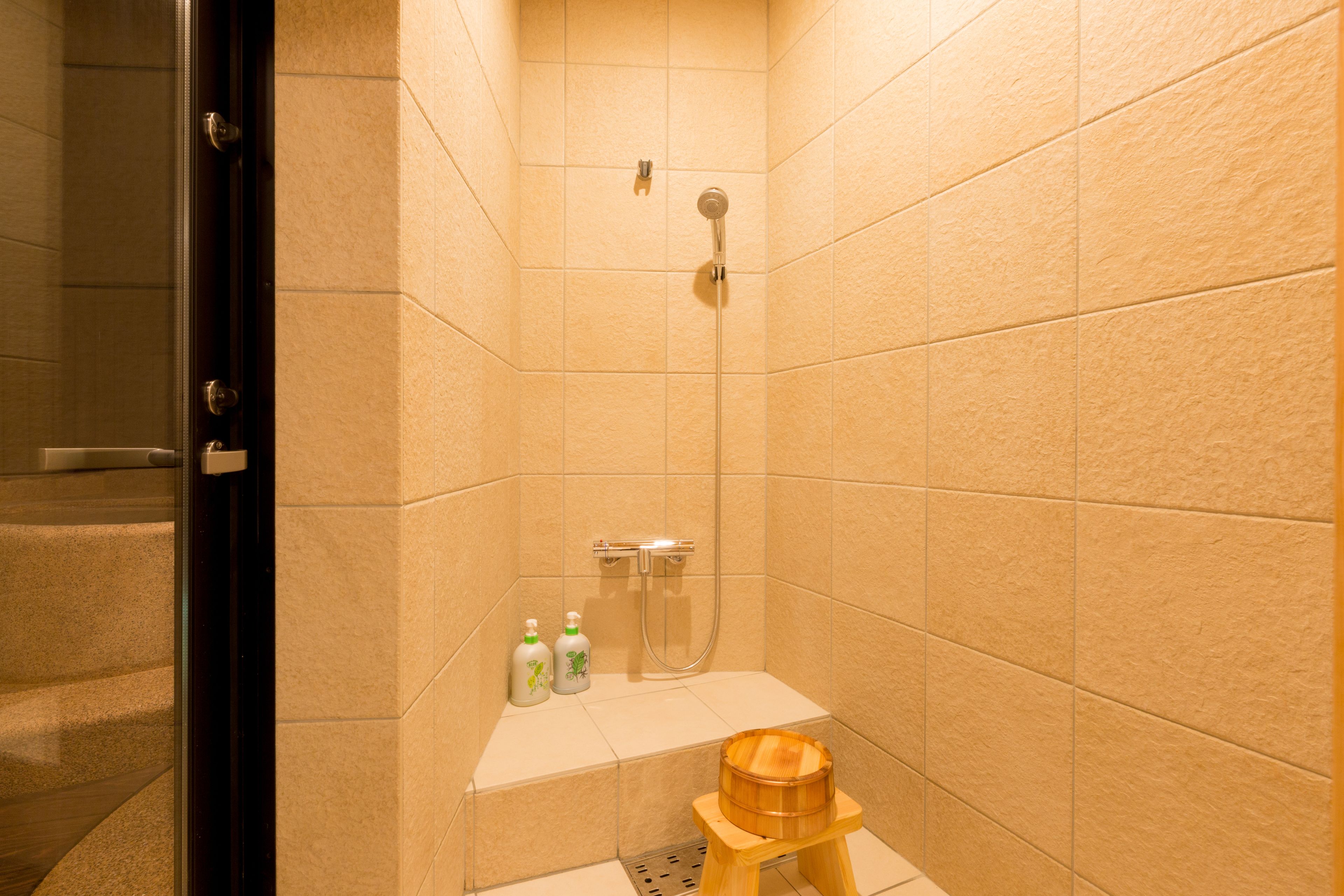 A small bathroom with beige tiles, a shower, a wooden stool, and two bottles of shampoo.