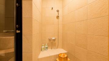 A small bathroom with beige tiles, a shower, a wooden stool, and two bottles of shampoo.