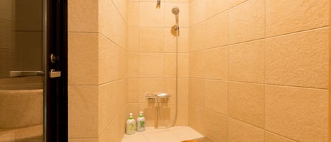 A small bathroom with beige tiles, a shower, a wooden stool, and two bottles of shampoo.