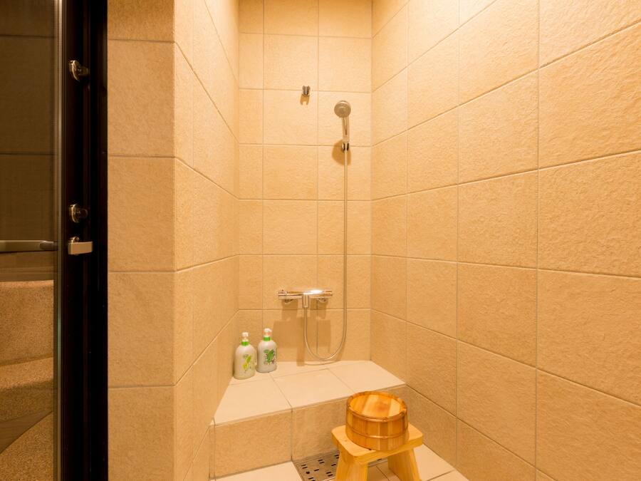 A small bathroom with beige tiles, a shower, a wooden stool, and two bottles of shampoo.