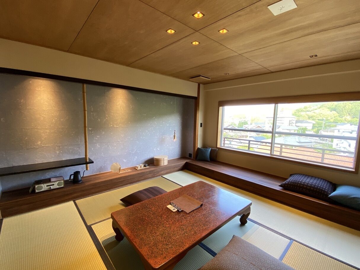 Una habitación tradicional japonesa con suelo de tatami, una mesita baja, cojines y vistas a través de un ventanal grande.
