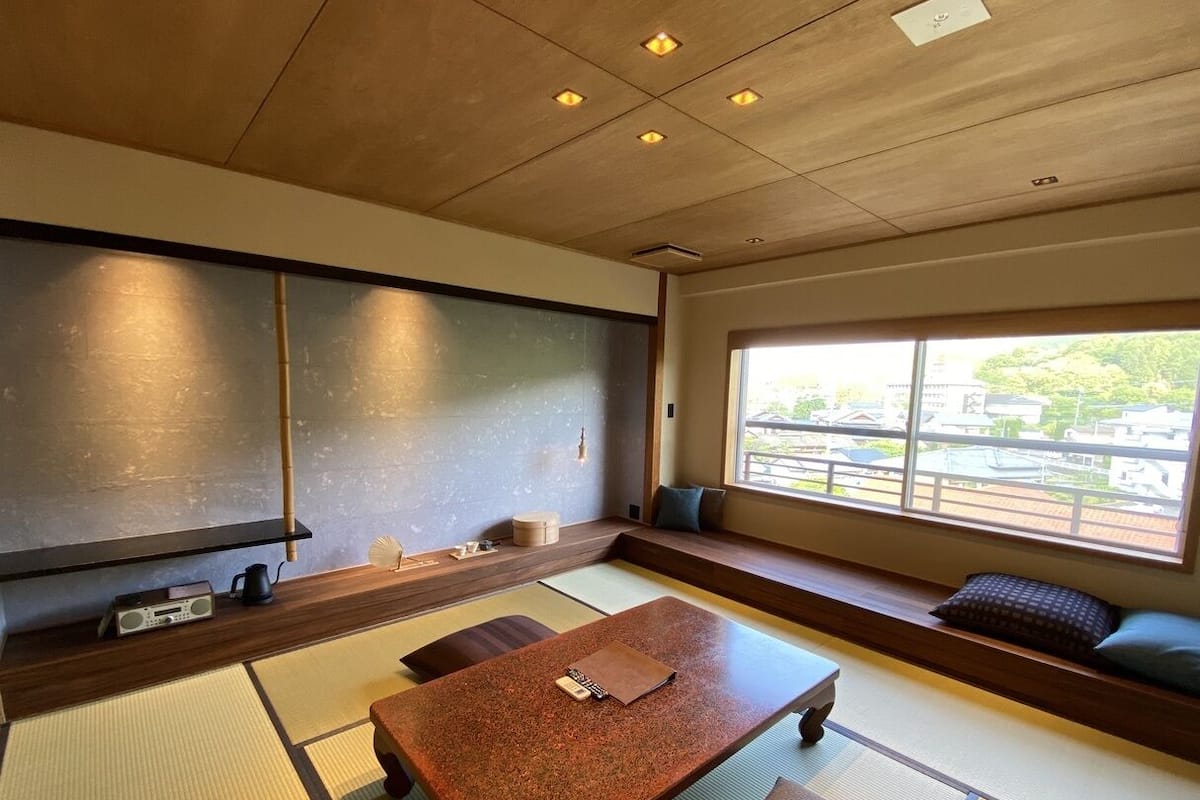A traditional Japanese-style room with tatami flooring, a low table, cushions, and a view through a large window.
