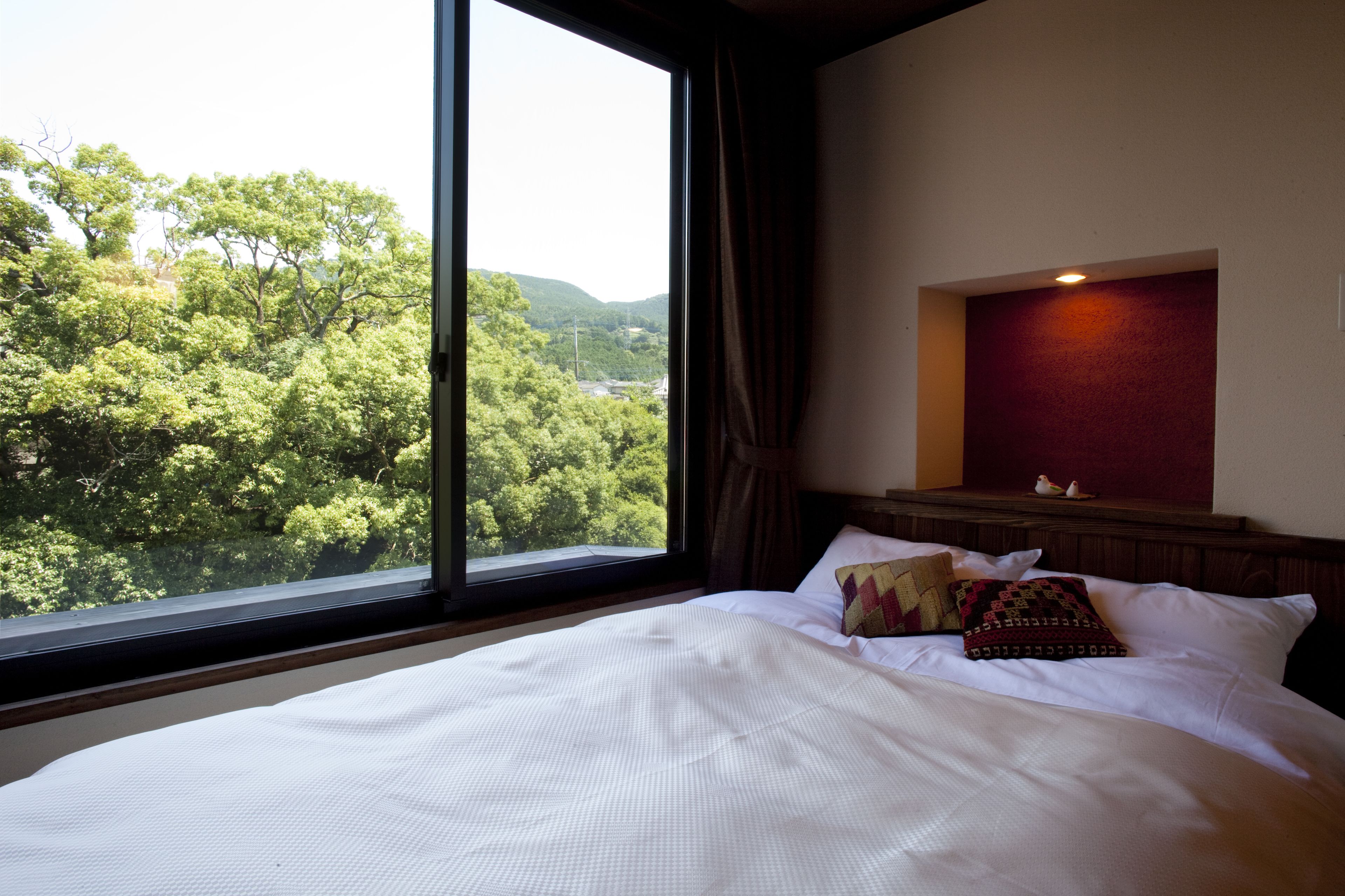 A hotel room with a large window, a bed with white linens, and a view of green trees and hills.