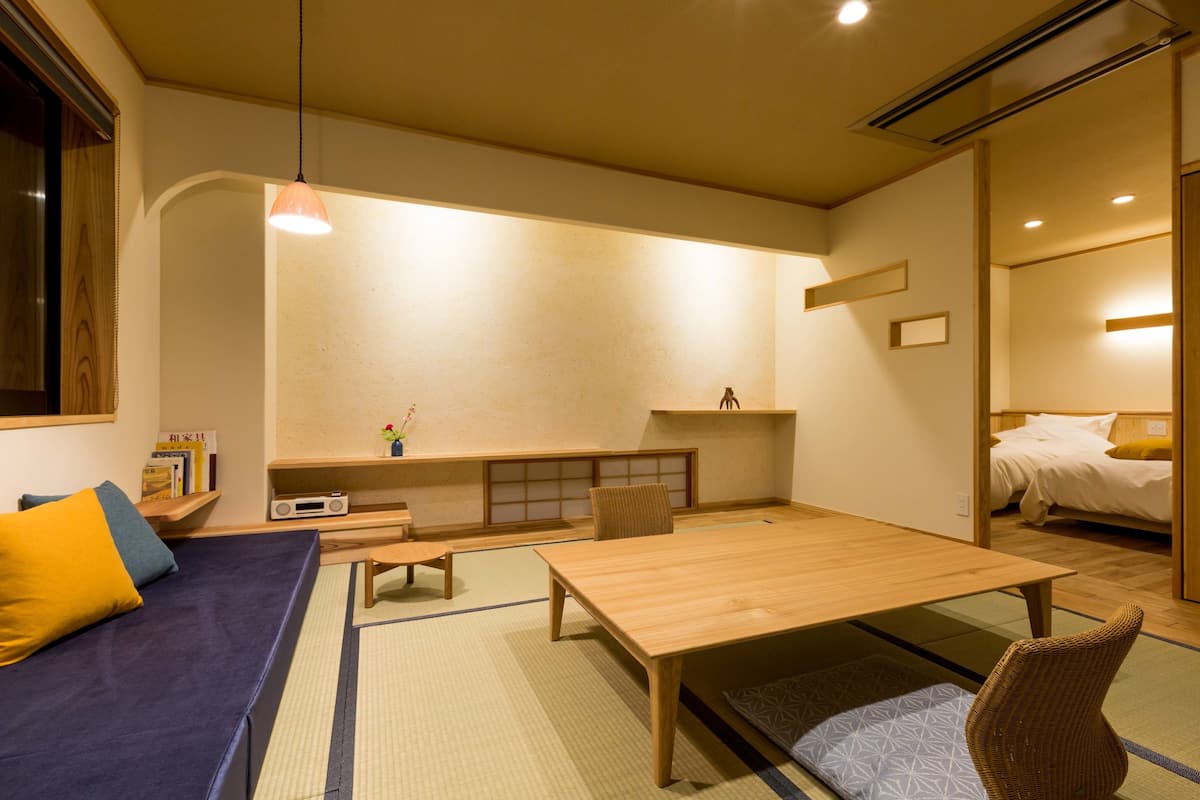 A traditional Japanese-style room with tatami flooring, a low wooden table, a sofa with cushions, a small shelf with books, and a bed in the background.