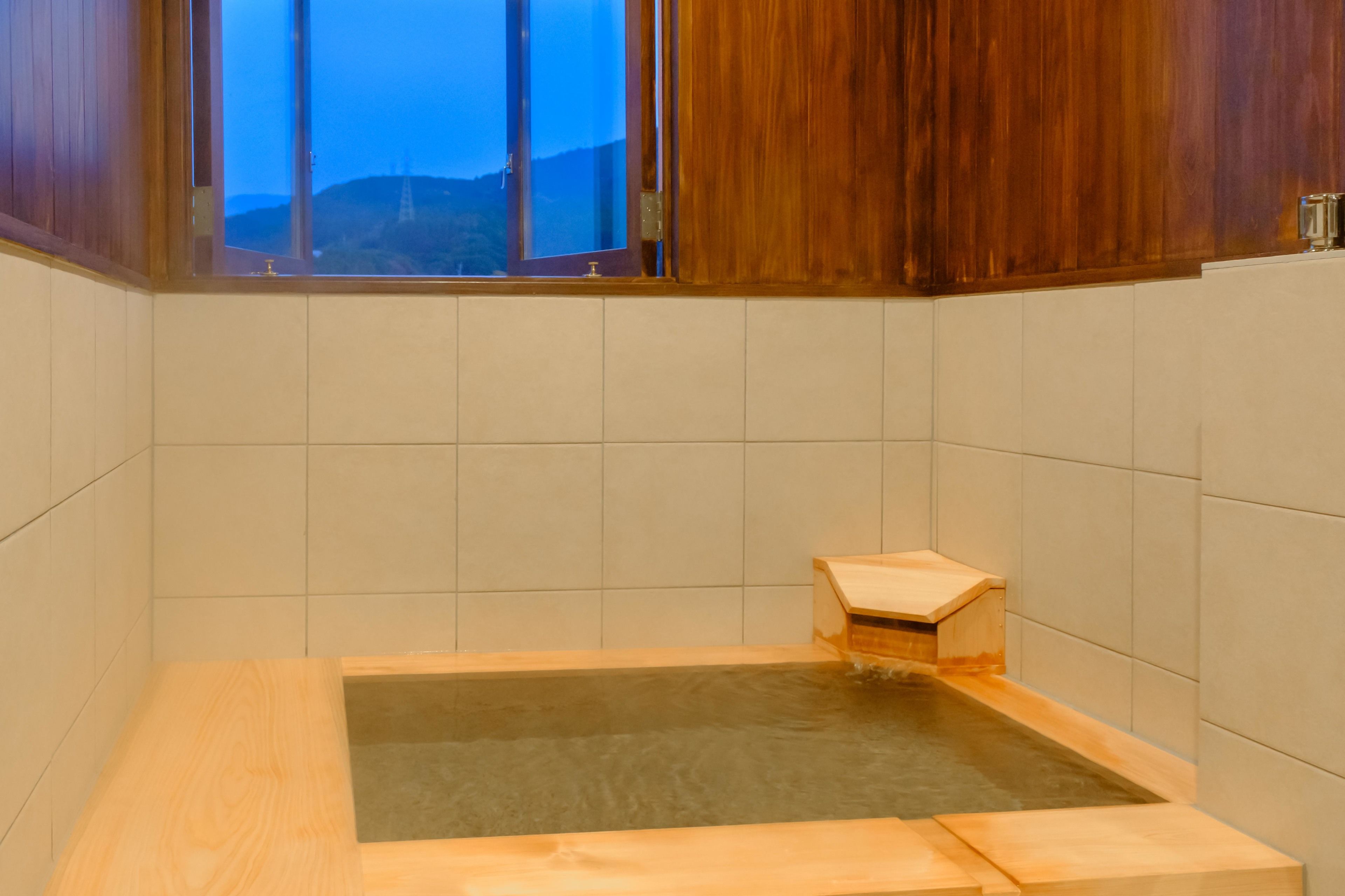 A wooden bathtub with a small wooden platform in a room with tiled walls and a window overlooking hills.