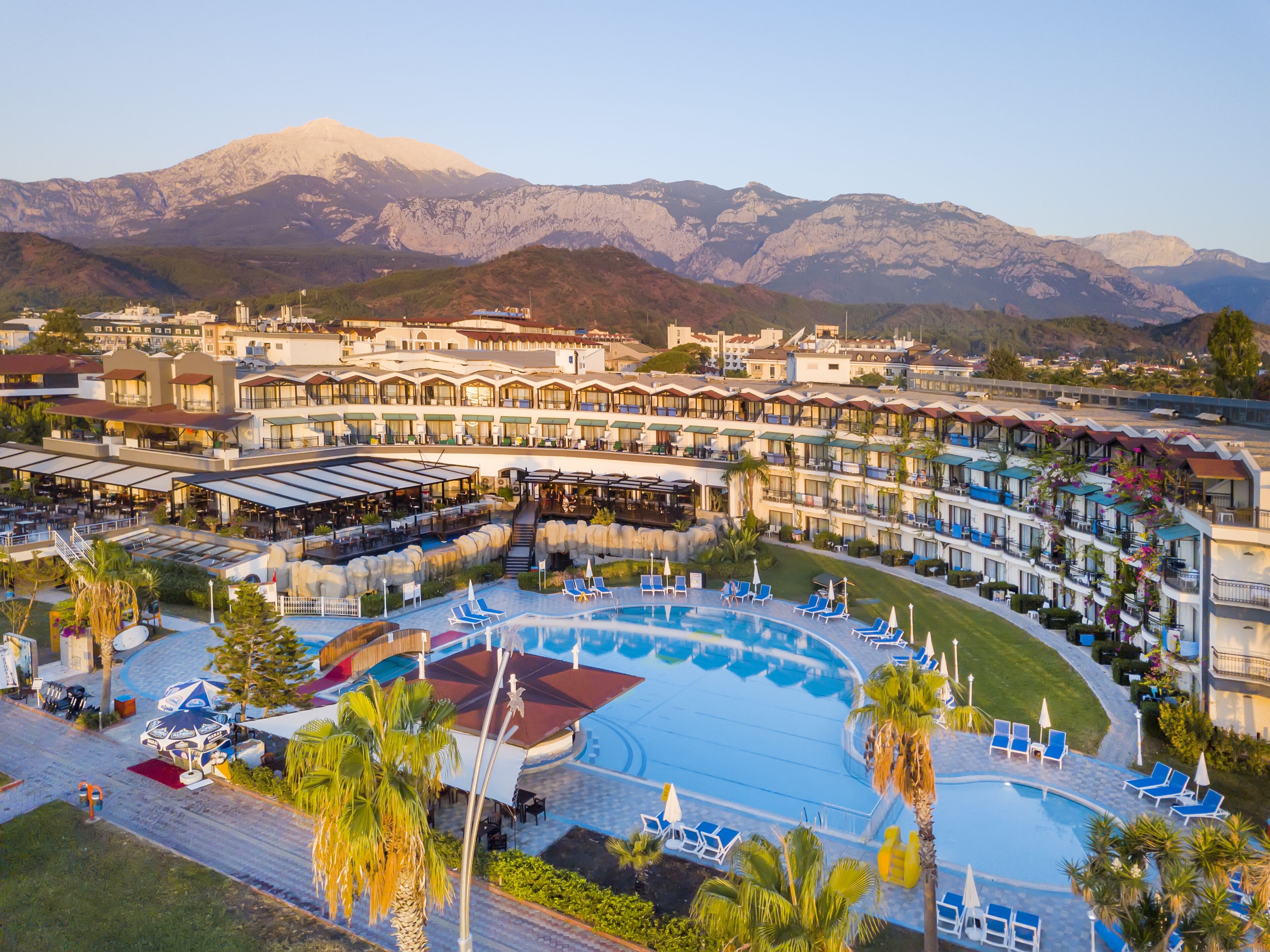 Un resort con una gran piscina, varias habitaciones para huéspedes y montañas al fondo.