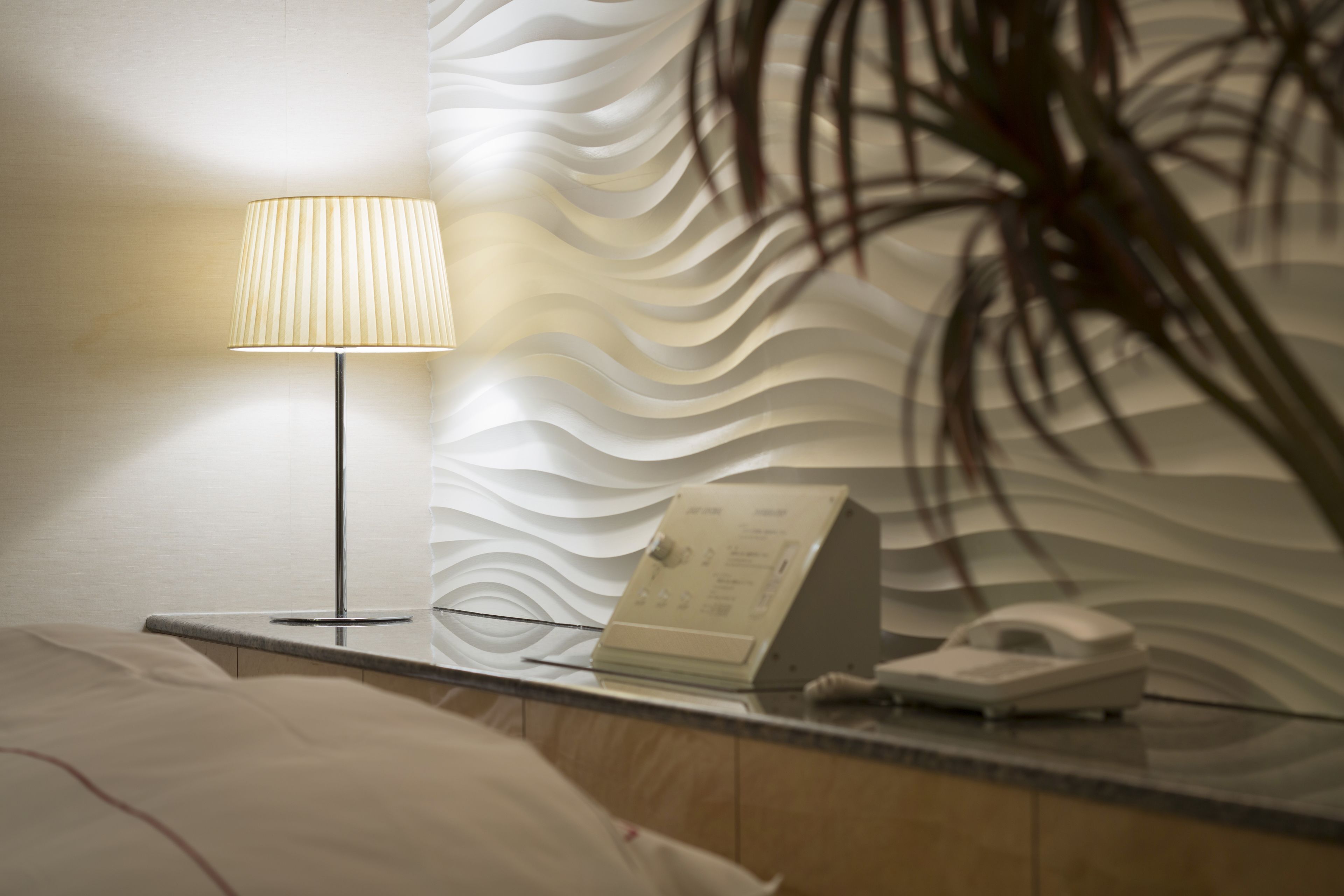 Hotellihuone, jossa on sänky, yöpöytä lampun kanssa, puhelin ja koristeellinen seinä.