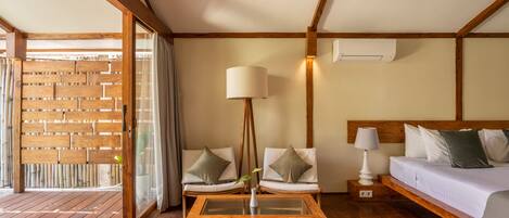 A room with a wooden floor, a bed with white bedding, two chairs with cushions, a small table with a lamp, and a wooden partition with a window.