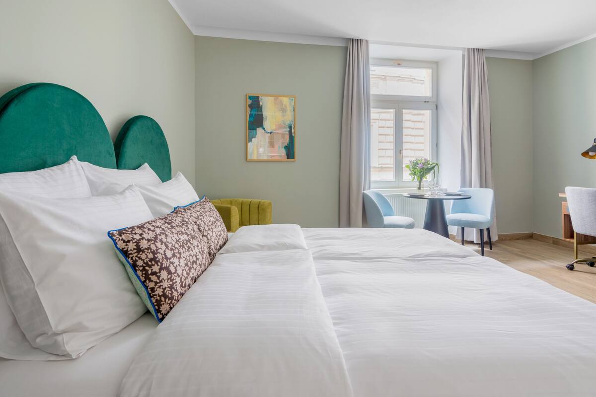Una cama bien hecha con sábanas blancas y almohadones decorativos, una pequeña mesa redonda con un jarrón de flores y una silla en una habitación con paredes verde claro y una ventana con cortinas.
