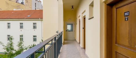 A corridor with a wooden door marked 202, a balcony with a metal railing, and a view of buildings with windows and a red roof.