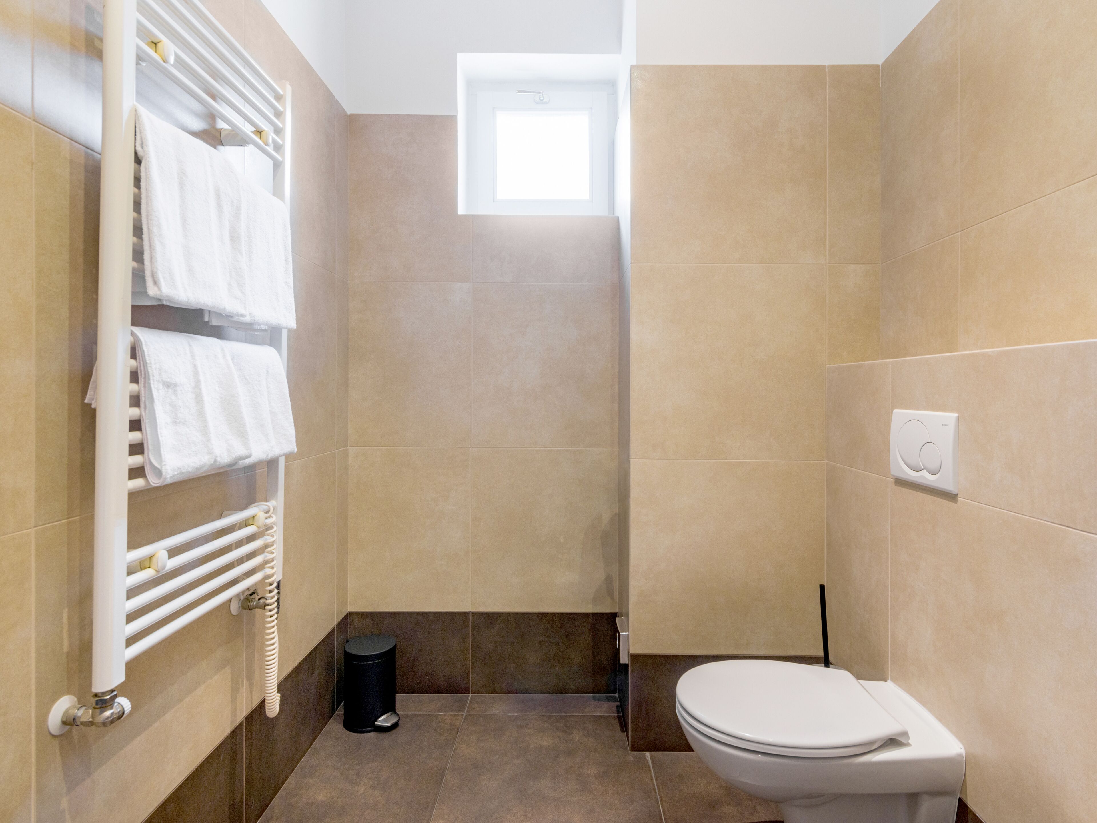 A compact bathroom with a toilet, a towel rack with folded towels, a black trash bin, and a small window providing natural light.