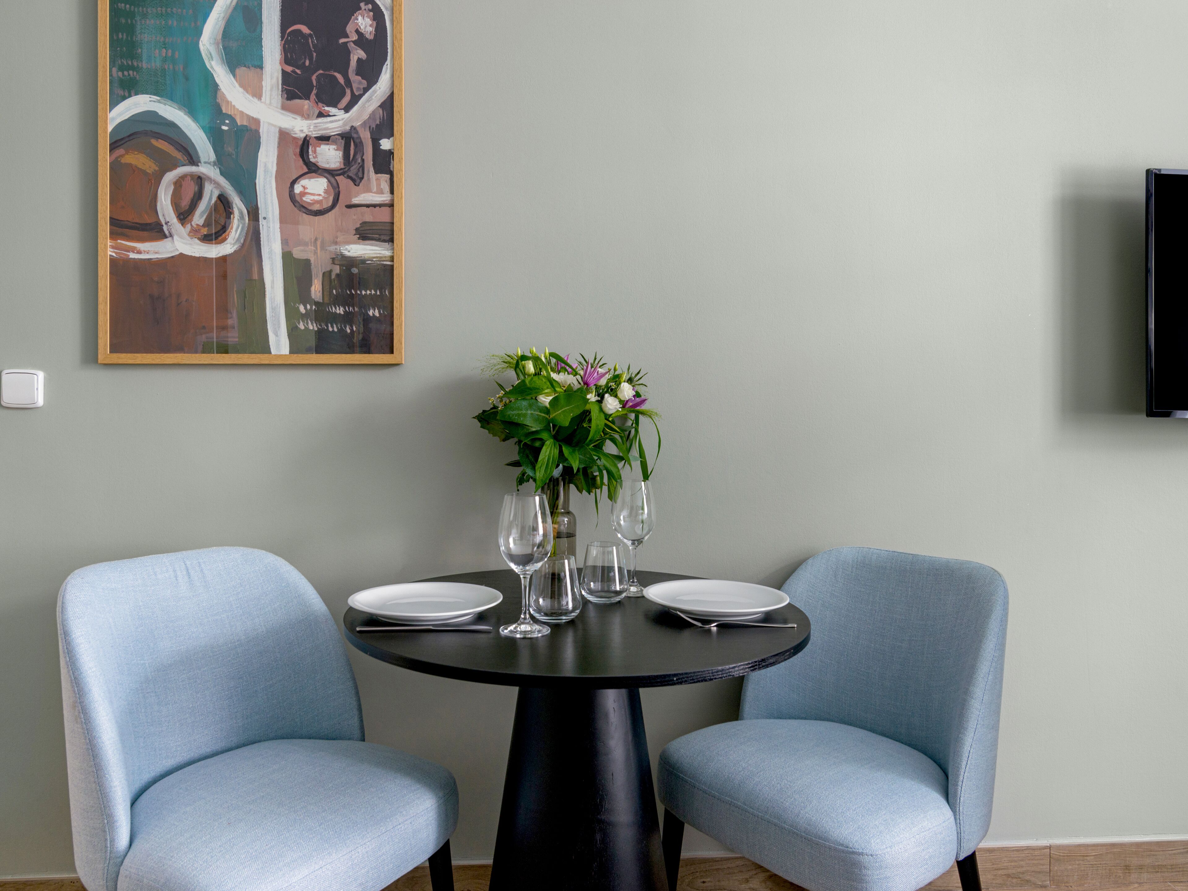 A dining area with two light blue upholstered chairs, a round black table, and a vase with flowers.