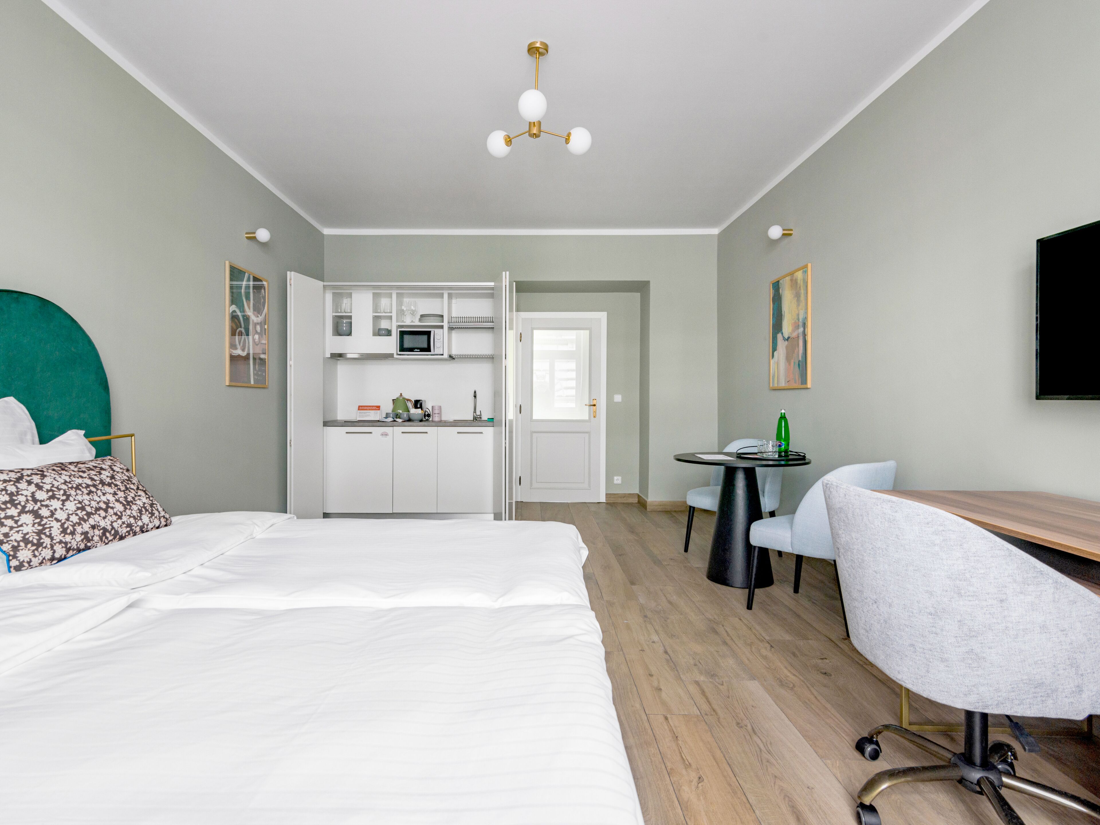 A hotel room with a bed, a desk, a chair, a kitchenette, and a TV.
