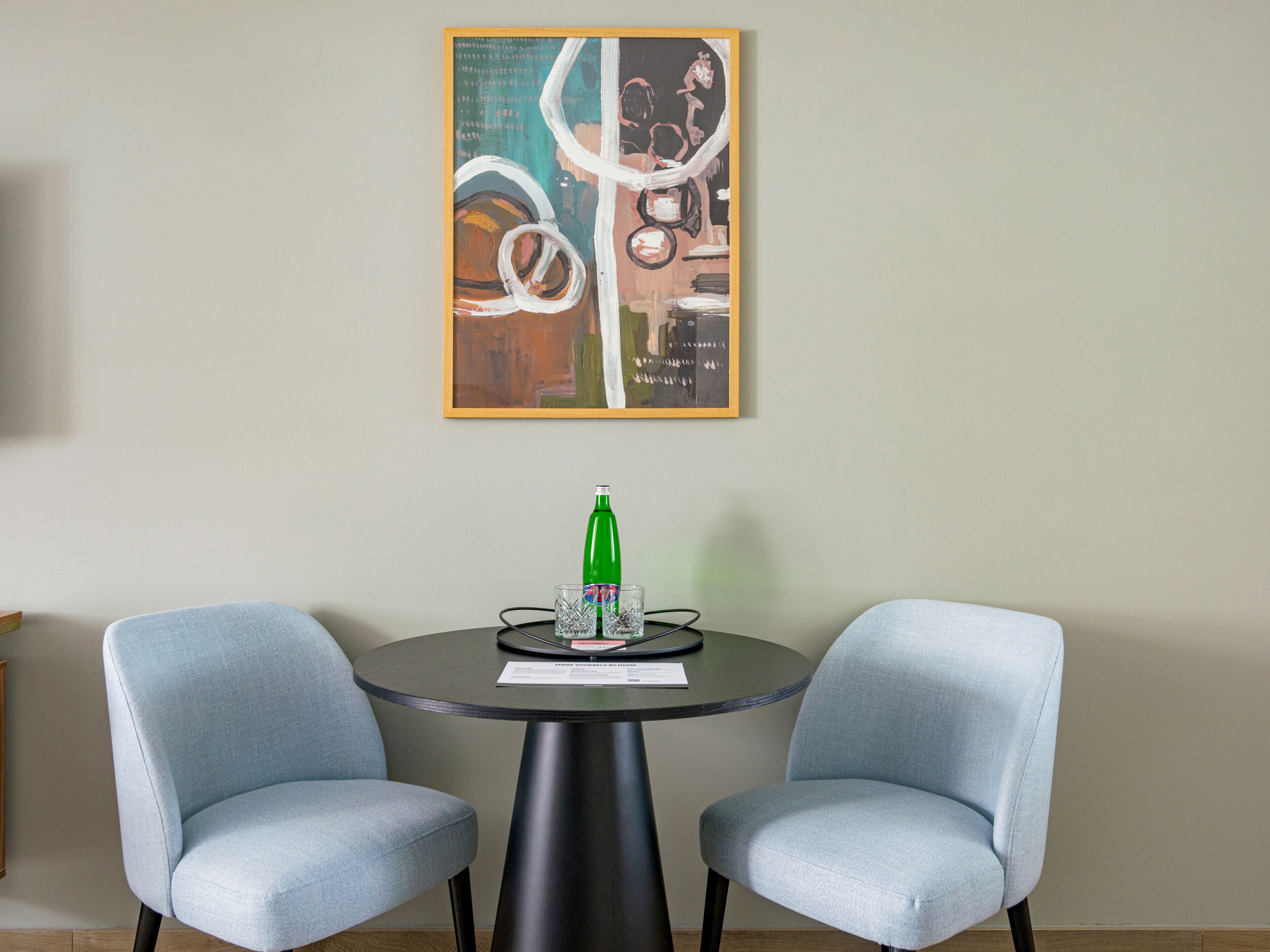 Een kamer met twee lichtblauwe stoelen rond een klein, rond tafeltje, een groene fles en een abstract schilderij in een lijst aan de muur.