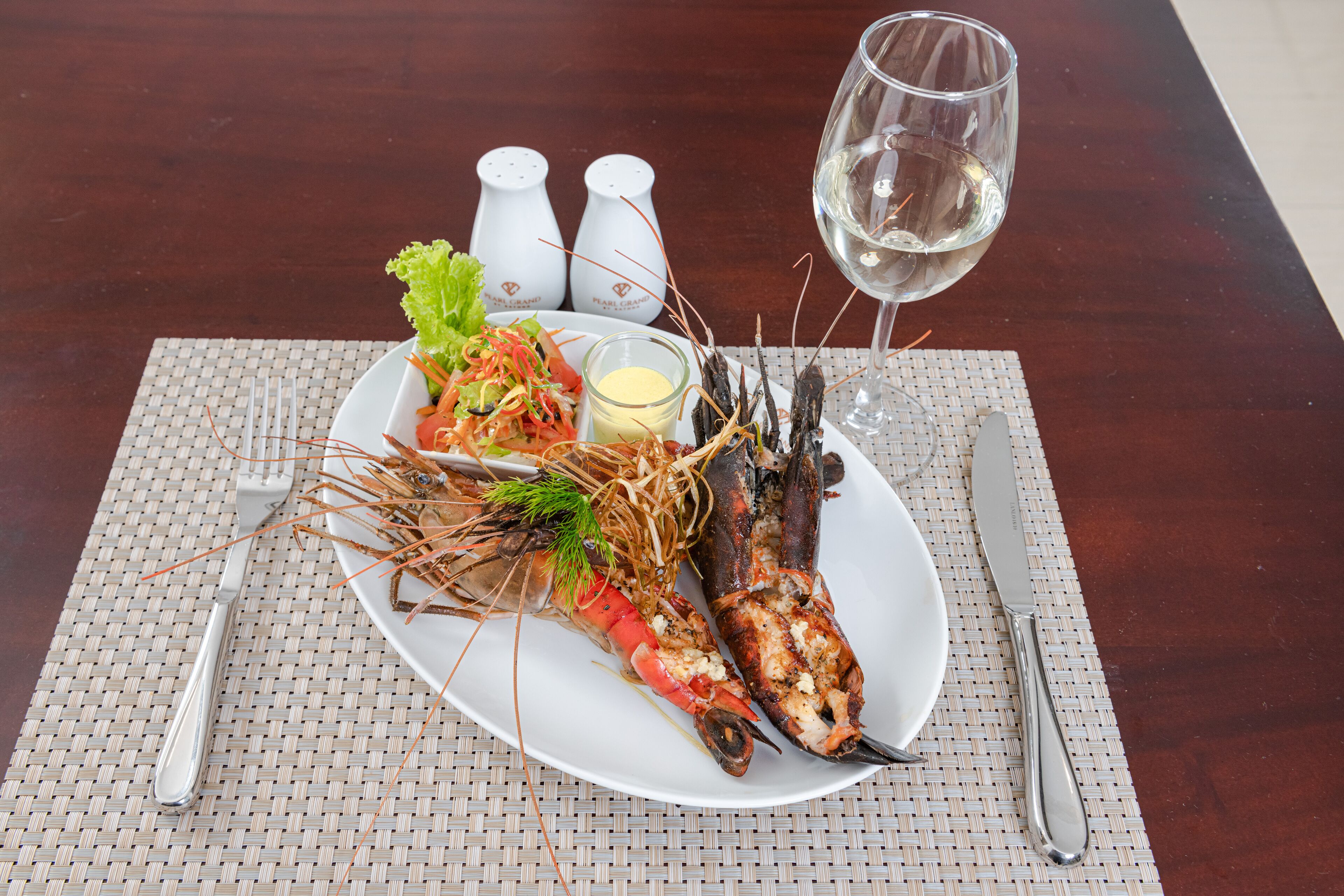 Une assiette avec du homard grillé et une salade, un verre de vin blanc, ainsi qu’une fourchette et un couteau posés sur un set de table en fibres tressées.