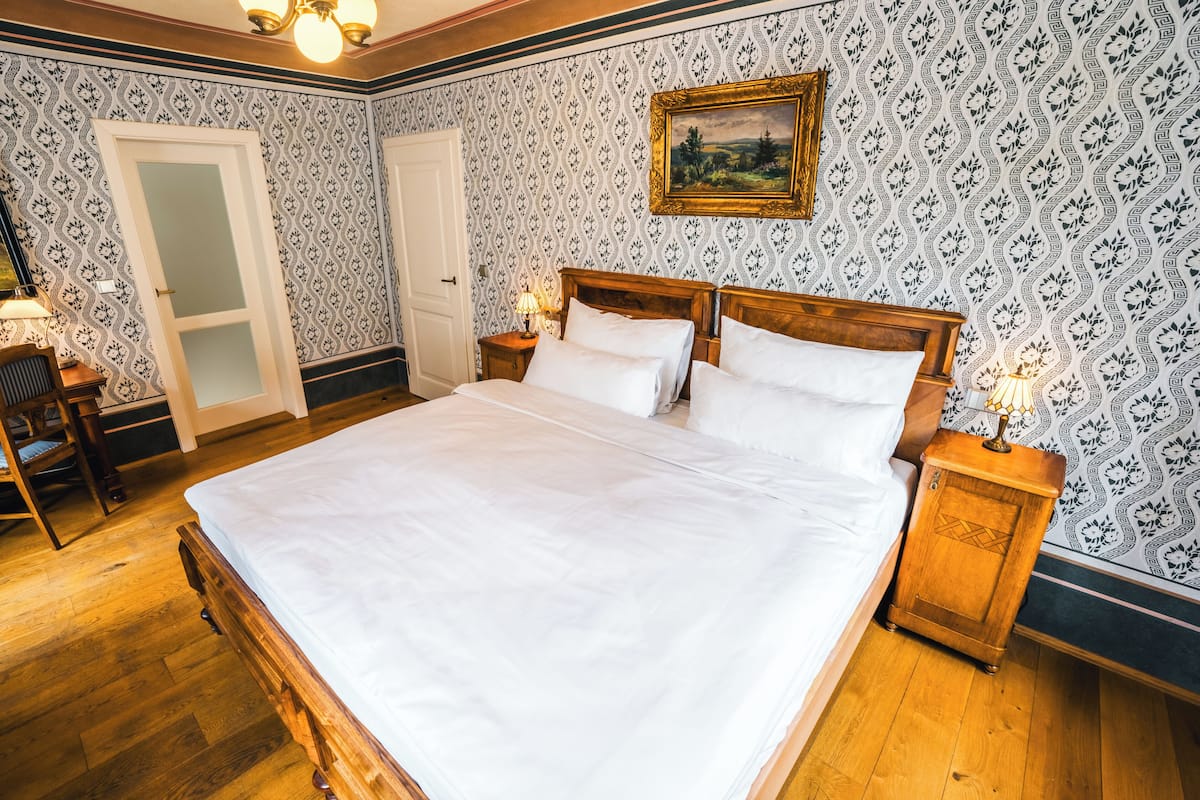 A neatly made bed with white linens in a room with patterned wallpaper and wooden flooring.