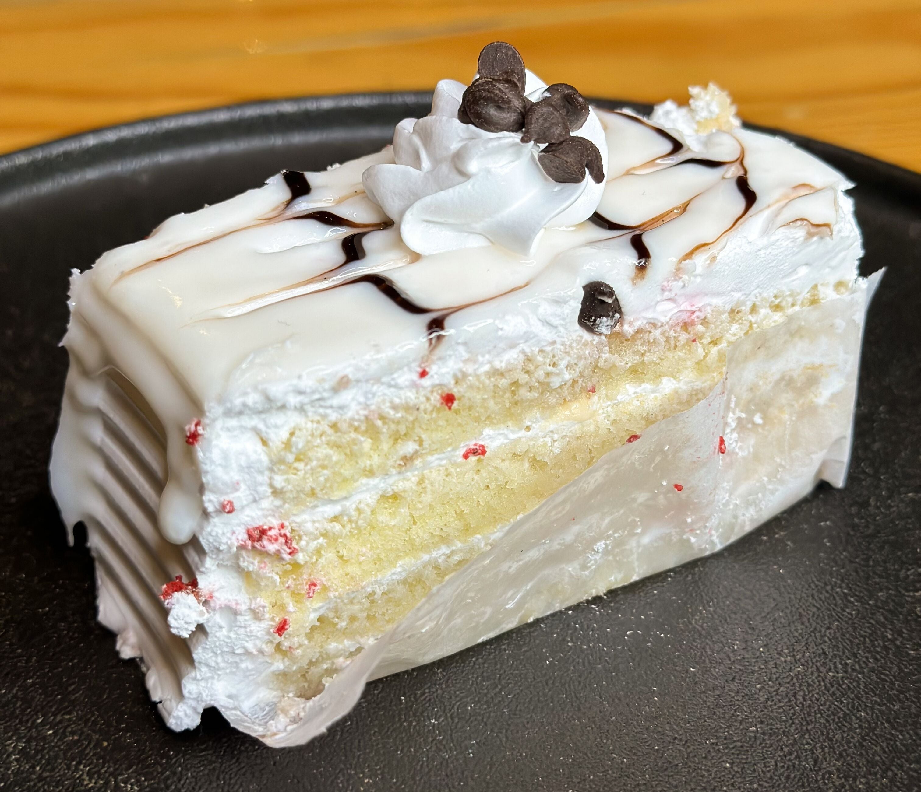 A slice of layered cake with white frosting, chocolate drizzle, and chocolate chips on a black plate.