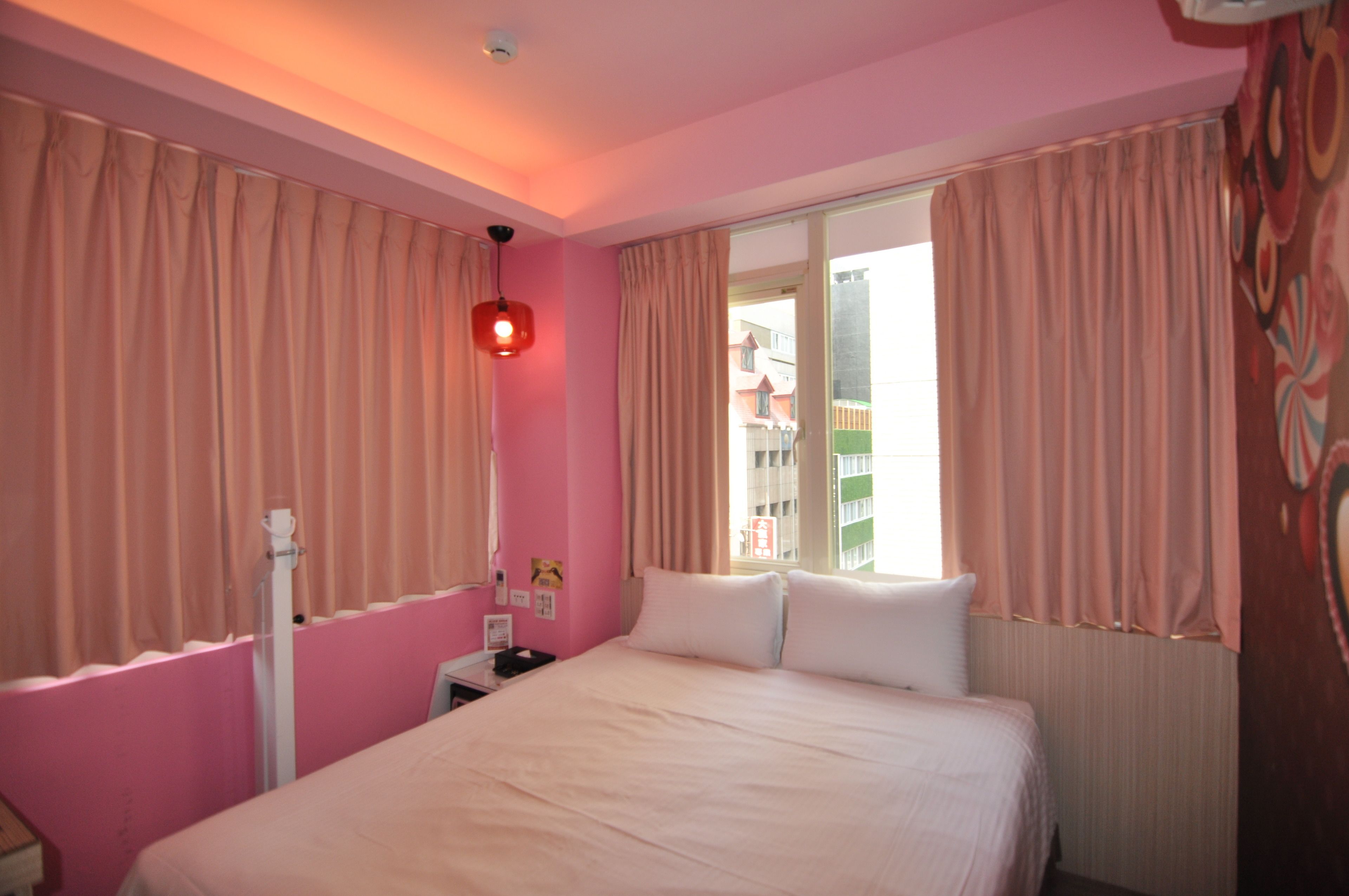 Una camera d'albergo con pareti rosa, un letto con lenzuola bianche, un comodino con una lampada e una finestra con tende.