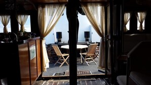 Ein Zimmer mit Holztisch und -stühlen, einer kleinen Küchenzeile und Meerblick durch transparente Vorhänge.