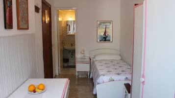 A small, well-lit room with a bed, a bedside table with a lamp, a chair, and a table with fruit.