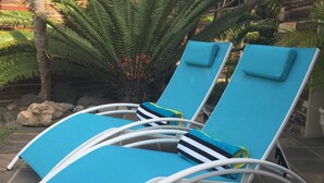 Dos sillones azules con estructura de metal blanco, ubicados en un patio de piedra con una planta de helecho y rocas al fondo.