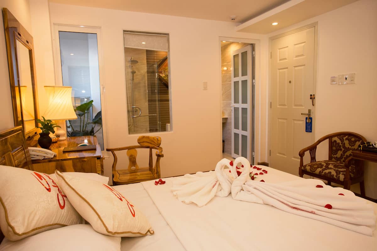 A hotel room with a bed, a chair, a desk with a lamp, a mirror, and a door.