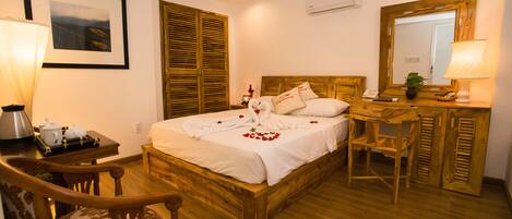 A neatly arranged bedroom with a wooden bed, a desk, a chair, and a bedside table with a lamp.