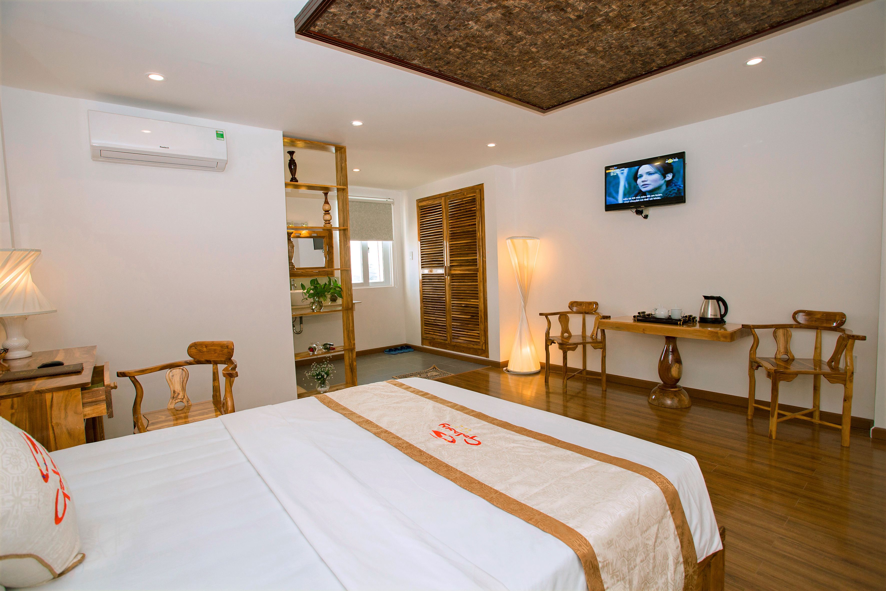 Habitación de hotel con cama, sillas de madera, televisor, mesa con hervidor y ventilador de techo.